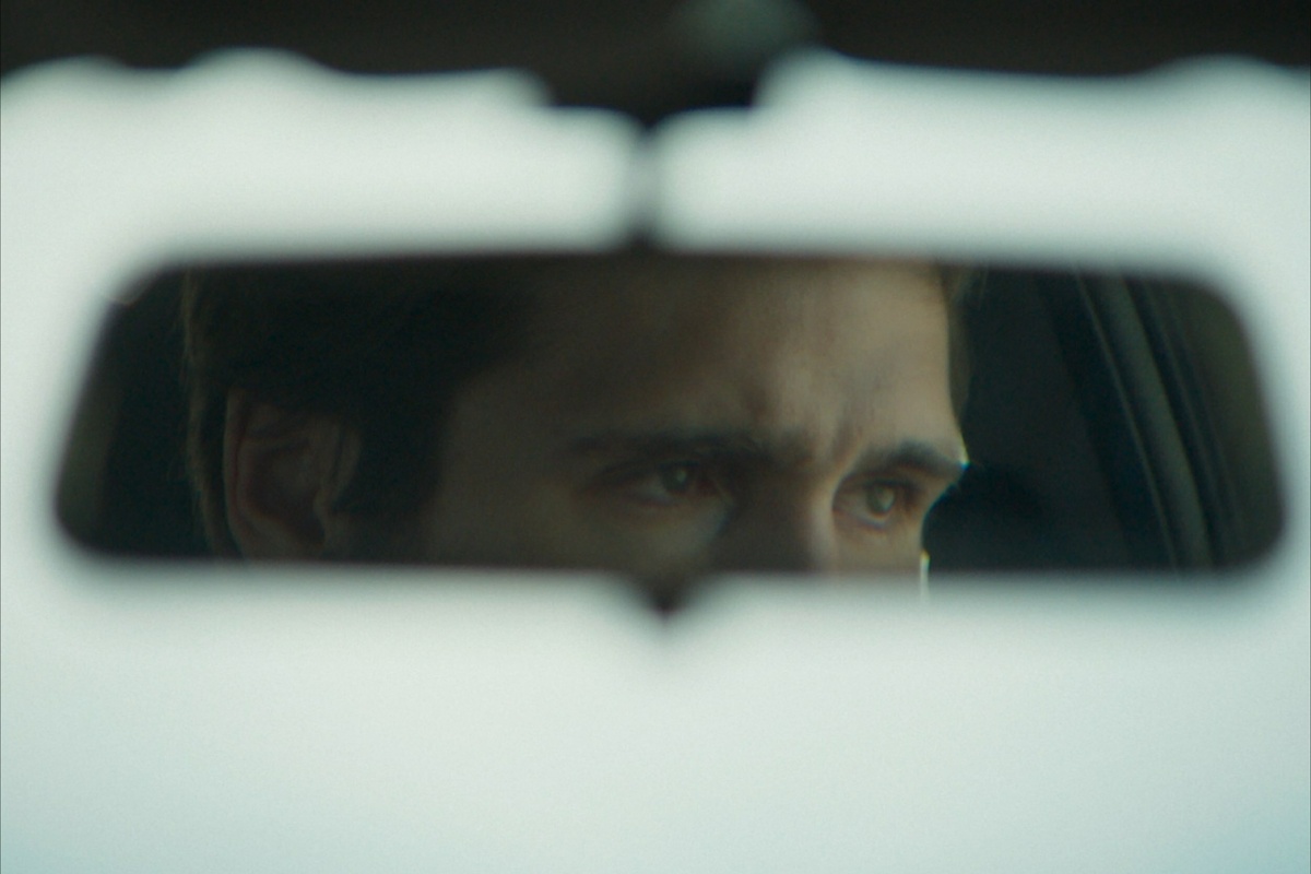 A rearview mirror of a car, showing the eyeline of a man’s face. He is looking to the right, concentrated and concerned.