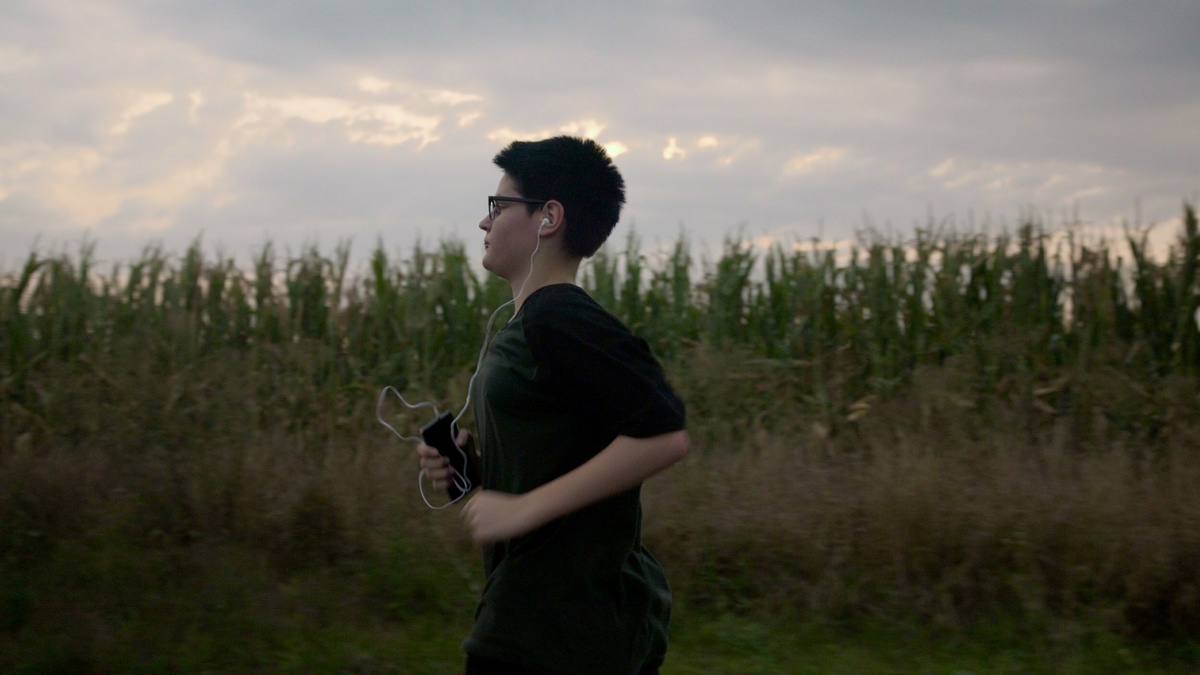 A person with dark hair and glasses is jogging in a field. They are carrying a music player and wearing headphones.