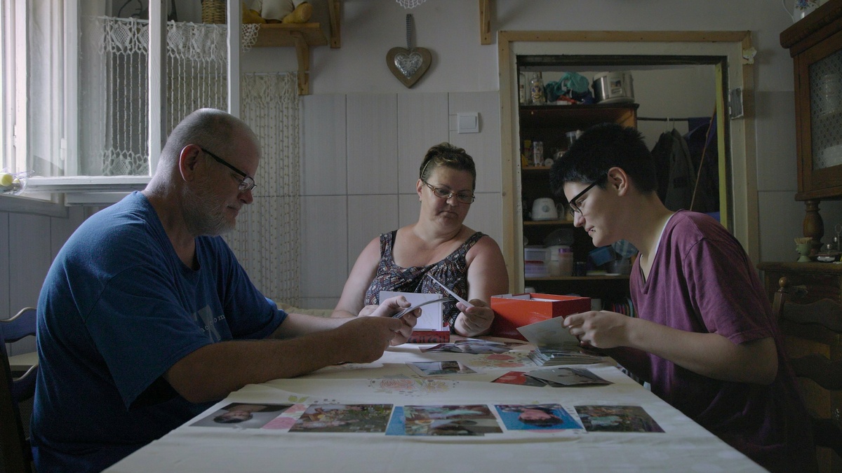 A middle-aged man, a middle-aged woman, and a teenager are sitting at a table and examining photographs laid out on it.