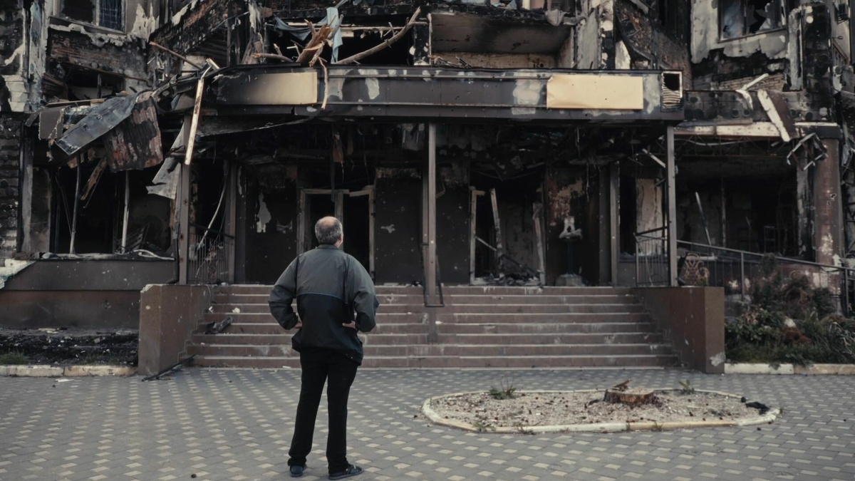 A man, with his back towards the screen, is looking at a building façade destroyed by russian missiles.