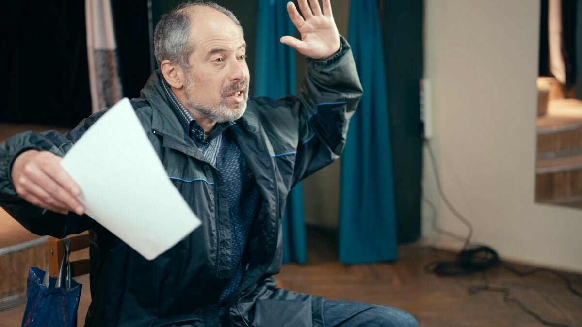 A white middle-aged man with receding grey hair is gesticulating while holding an A4 sheet of paper in his right hand.