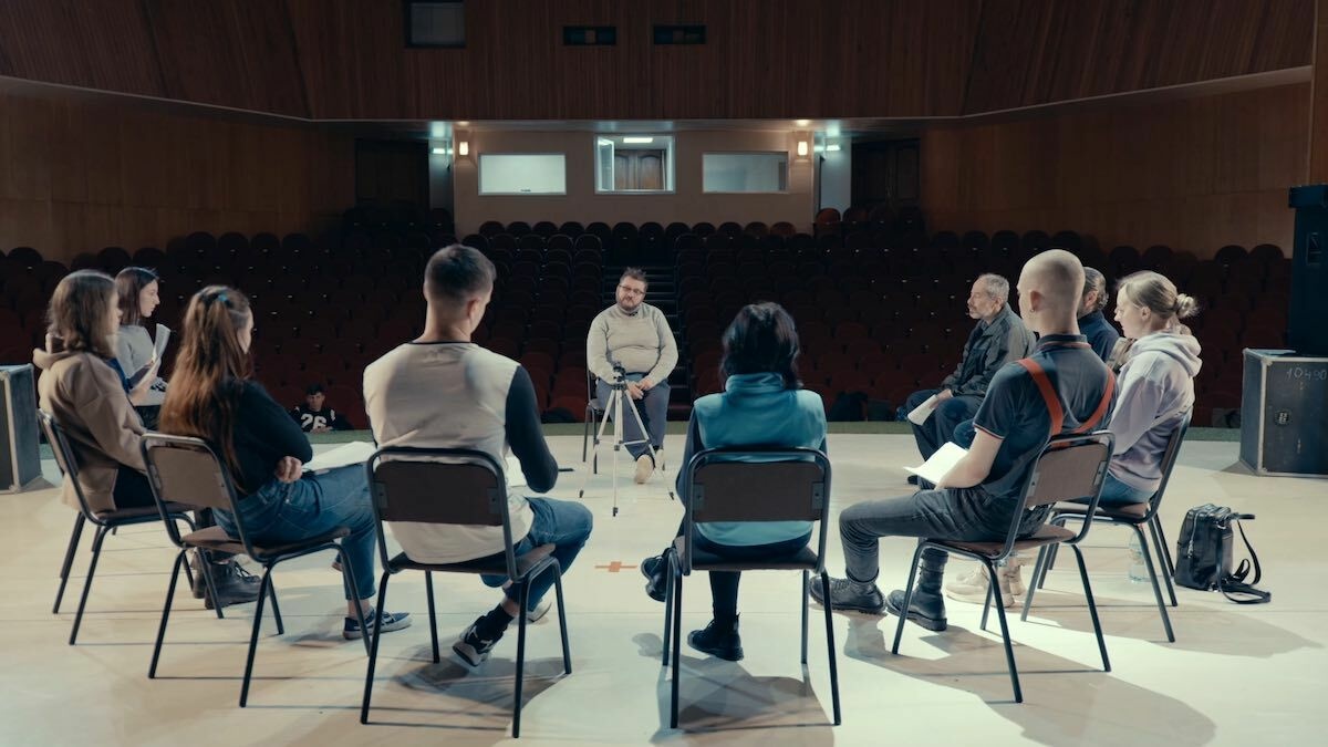 An empty theatre, on the stage of which we see several actors sitting in a semi-circle turned towards a man sweater.