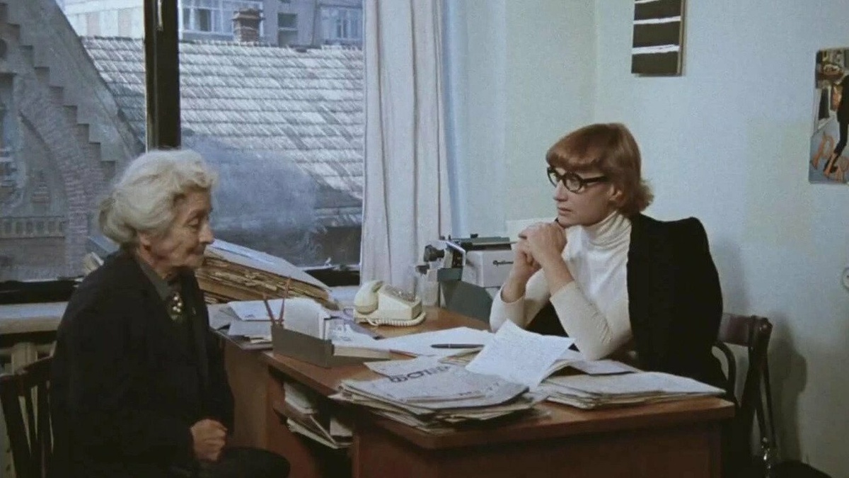 A middle-aged woman with ginger hair is sat behind a desk covered in papers. Opposite her is a older, grey-haired woman.