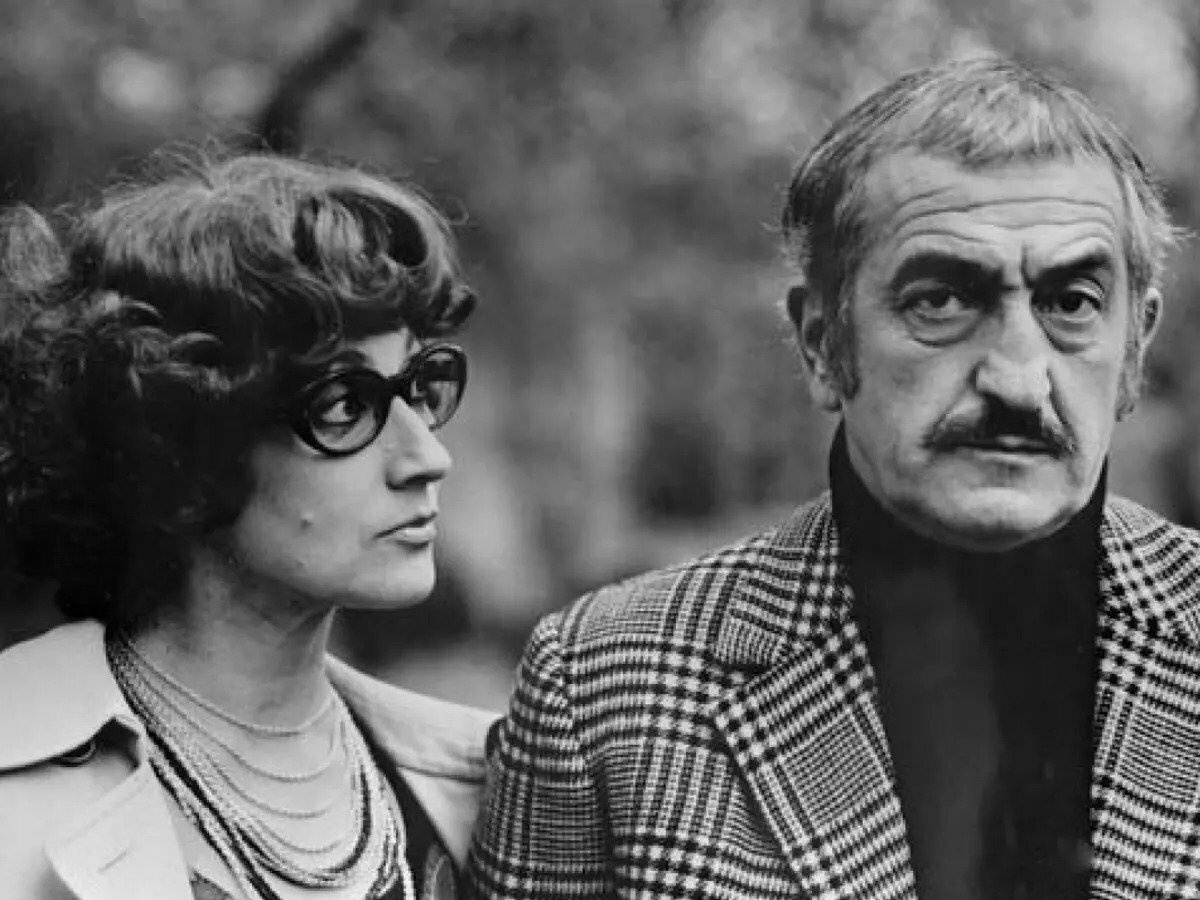 A black-and-white photo of a man and a woman. She has curly hair and glasses. He has a stern face, moustache, and blazer