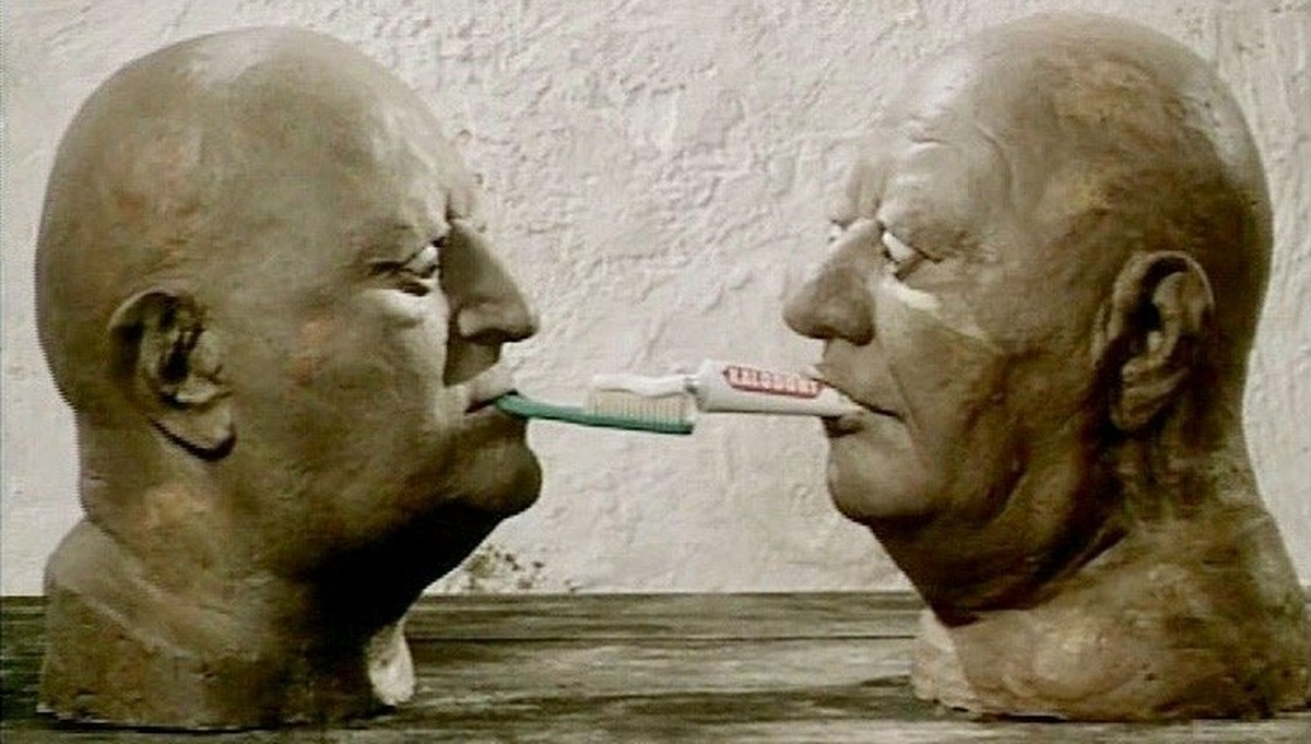 Two clay busts of bald men are standing on top of a table, facing each other with concentrated facial expressions.