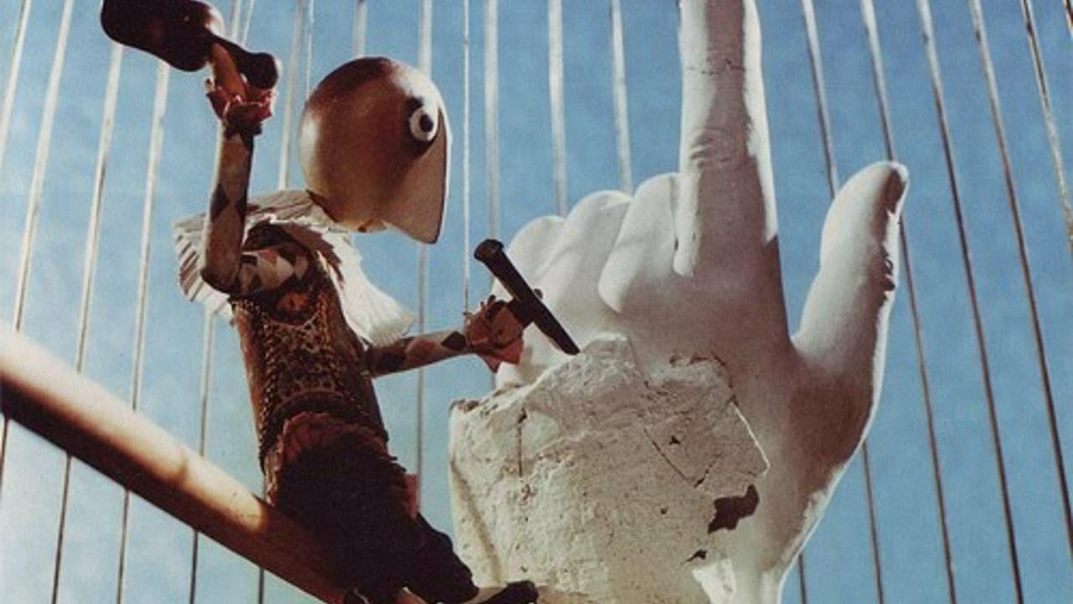 A puppet person is sitting in a birdcage and chiseling at a sculpture of a large human hand pointing to the sky.