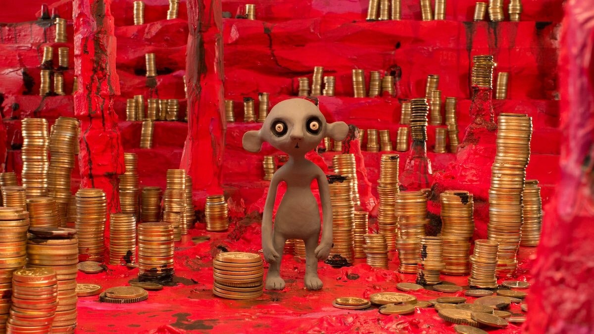 A grey clay mouse with sunken eyes is standing in a red cave, surrounded by stalagmites and stacks of coins.