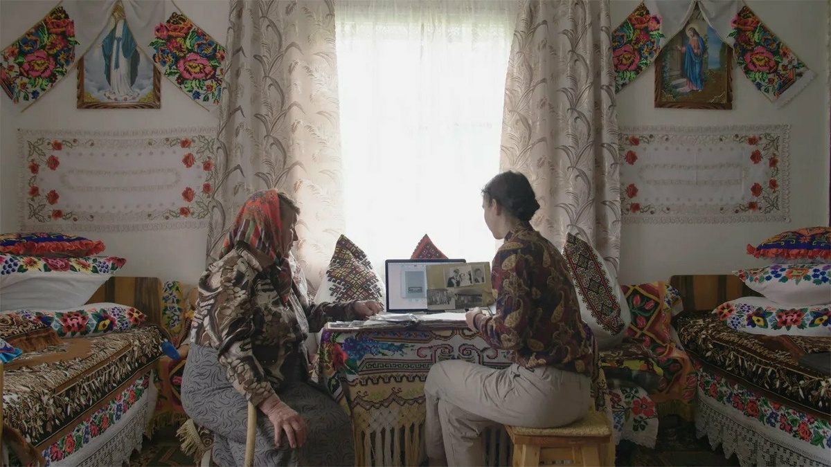 A young woman and a senior lady are sat by a table and looking at photos in a traditionally decorated Ukrainian house.