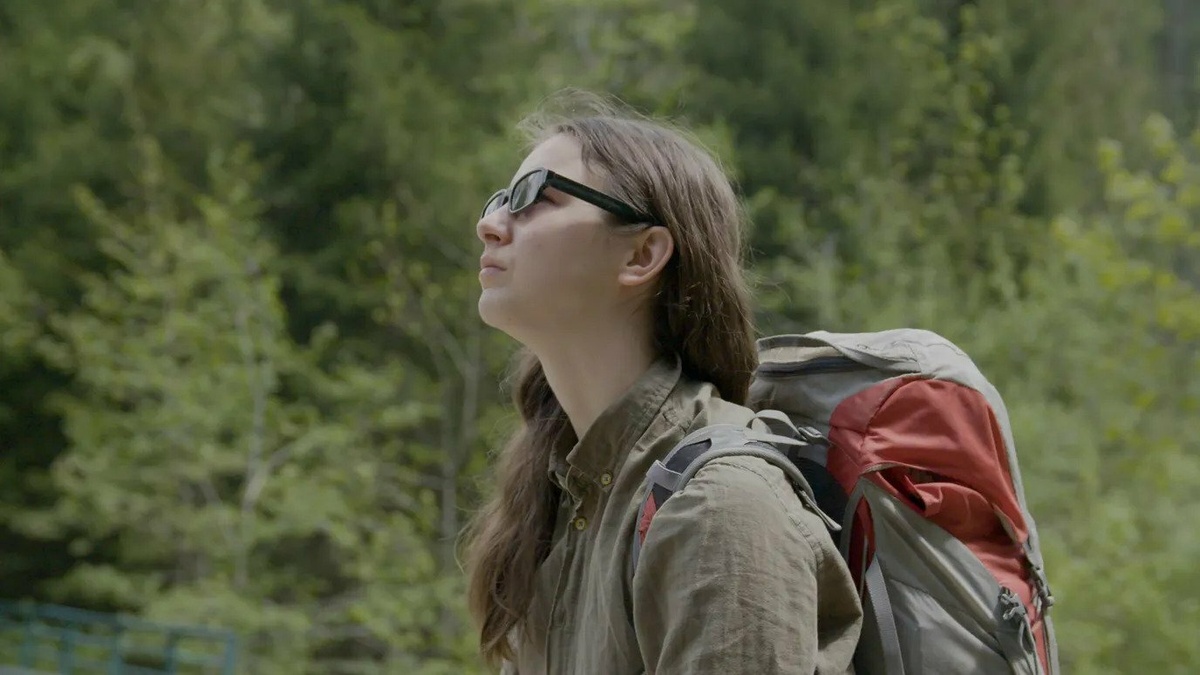A young woman with sunglasses and a backpack is looking up while walking in a forest area.