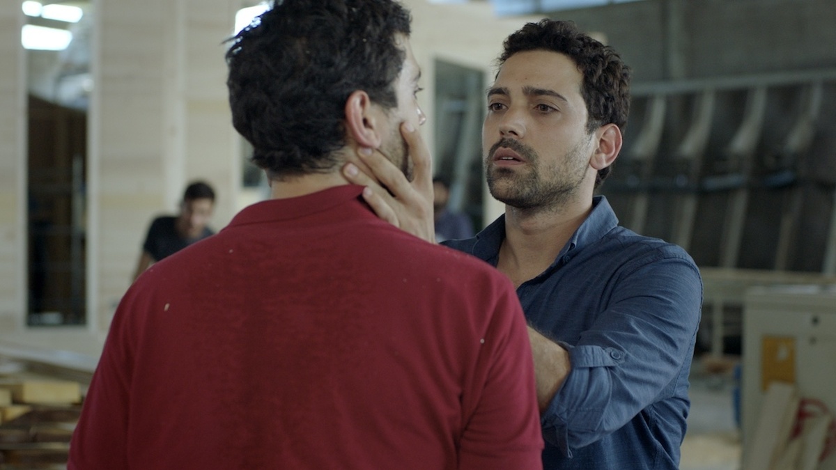 2 dark-haired male twins are looking at each other. The 1  in a blue shirt is holding the face of the 1  in a red shirt