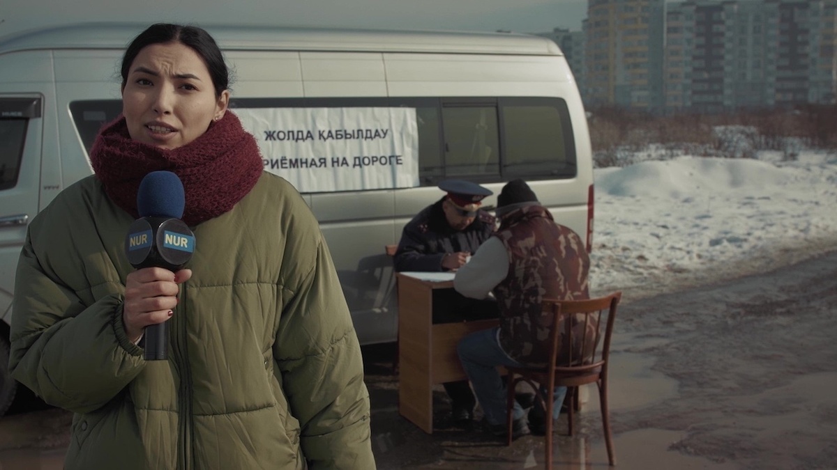 A female reporter in a winter coat with a mic by a van: ‘appointments on the street’ written in Russian & Kazakh