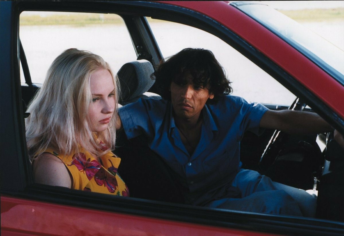 Inside a red car, a man with curly dark hair is turned towards a blond woman looking ahead.