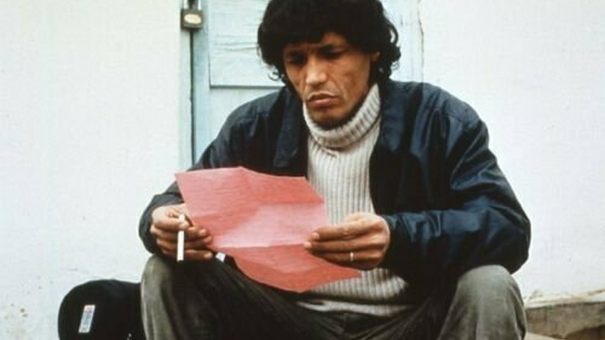 A curly man wearing a white sweater and black jacket is reading a pink letter as he’s smoking a cigarette.