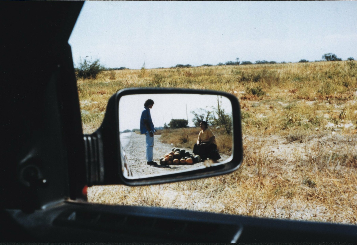 In a car’s side mirror, a reflection of a man buying a watermelon from a roadside seller.