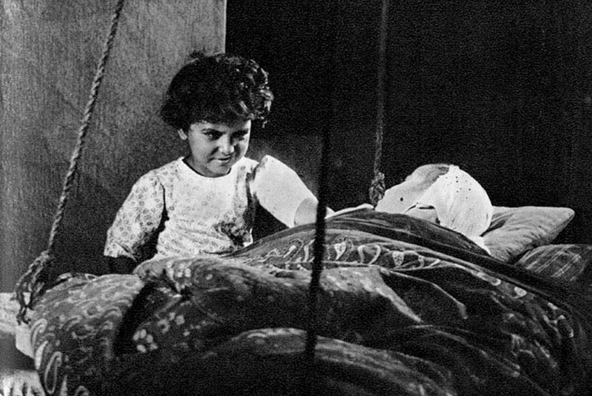 A curly-haired child is looking at another child in a hanging cot.