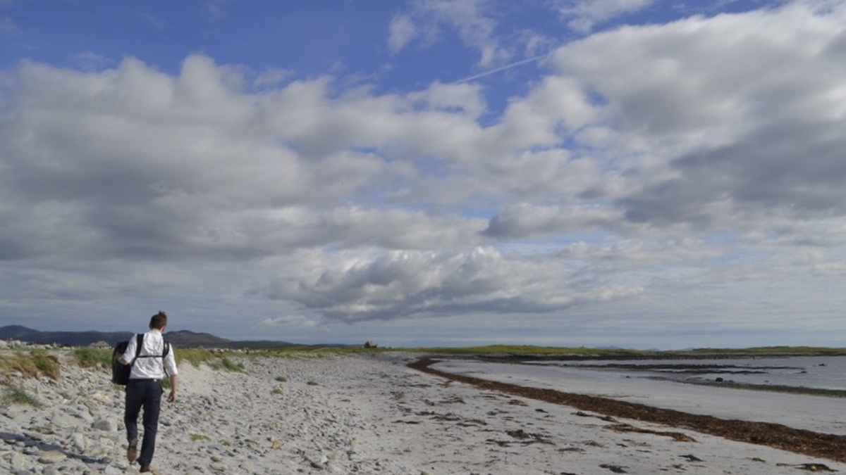 A lone figure carrying an accordion is walking into the distance on a sunny Hebridean beach.