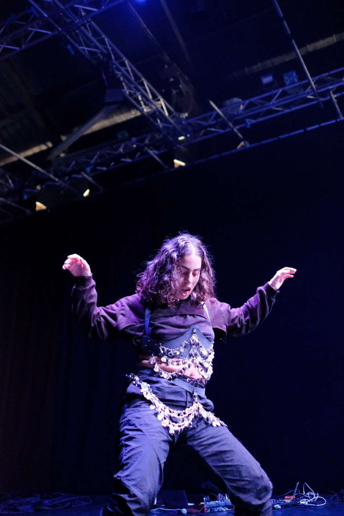 A person is performing within a dark theatrical space, they look down at the ground and holding their arms up mid-dance.
