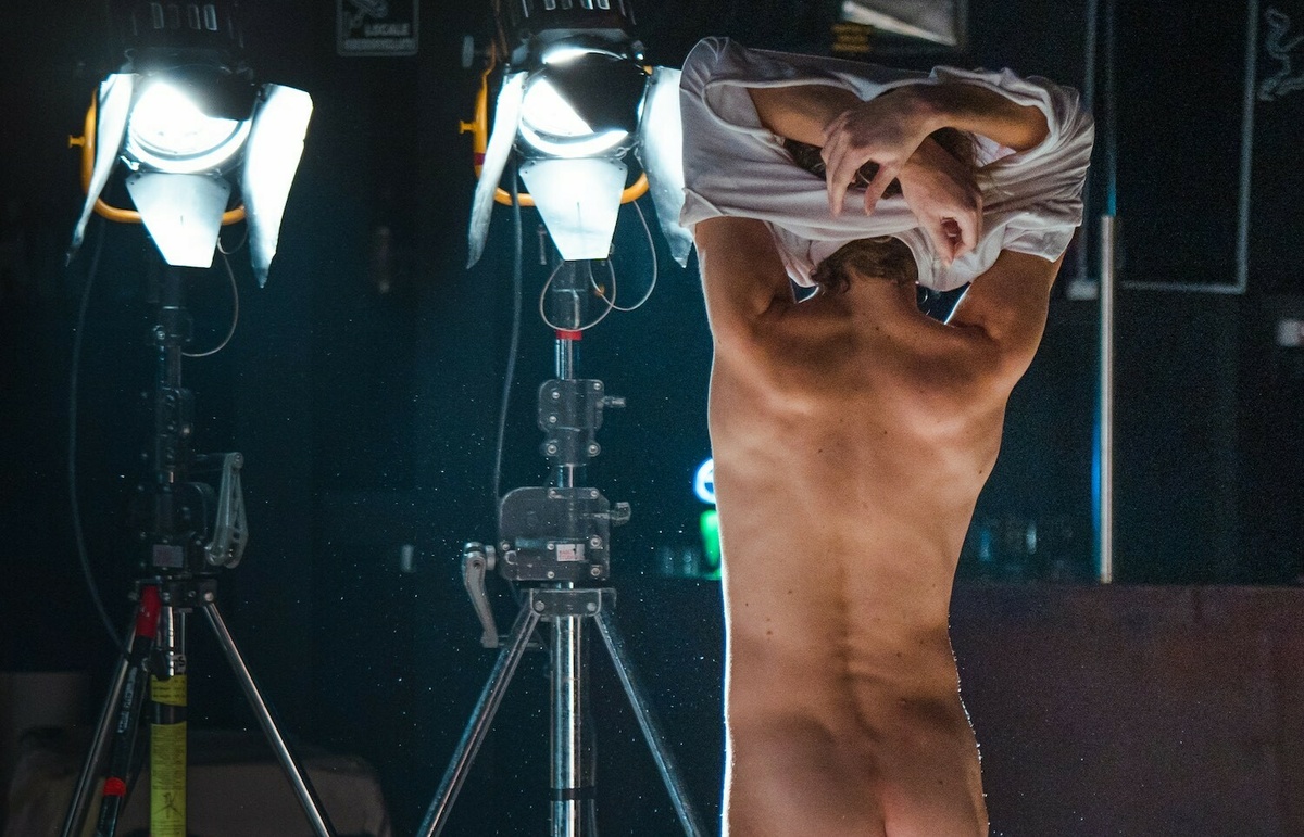 A person is facing two bright theatrical lights, they are naked and have lifted their clothing up over their head.