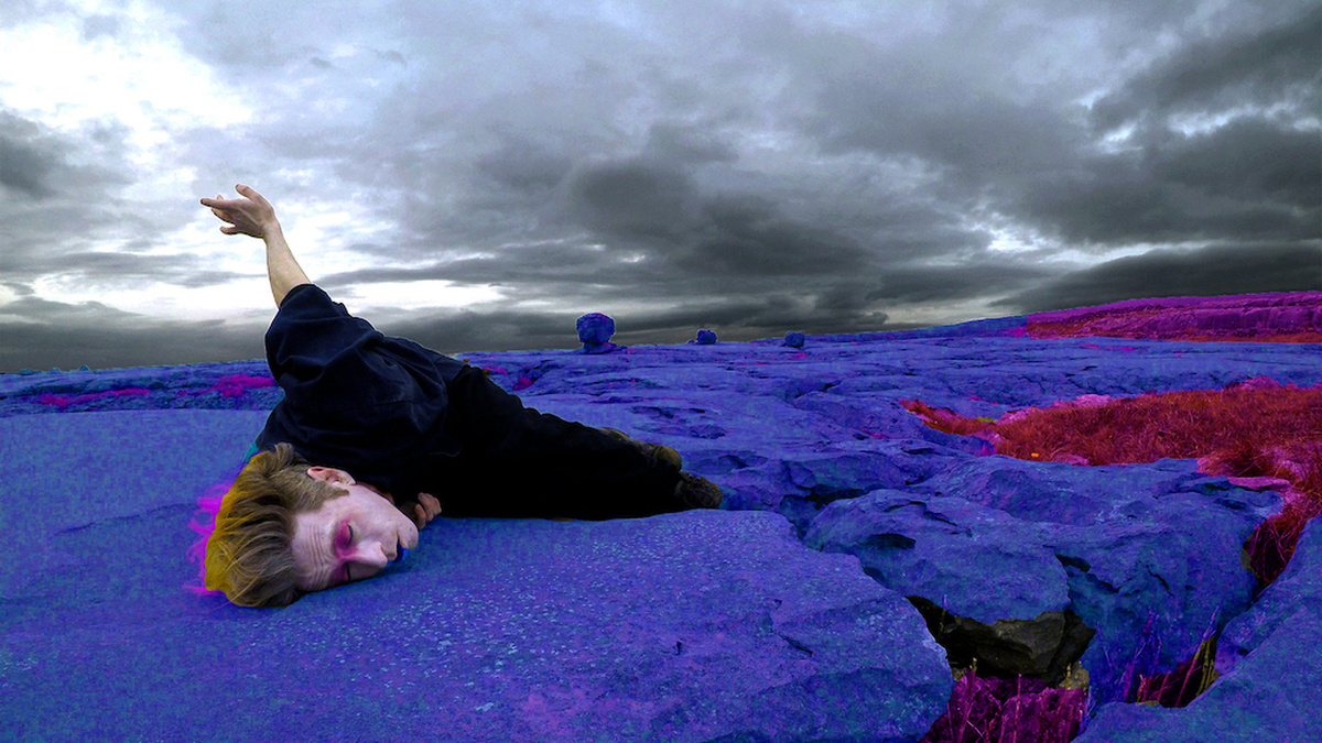 A dancer in a bright purple, rocky landscape, lying amongst the stones with one arm raised towards the stormy sky.