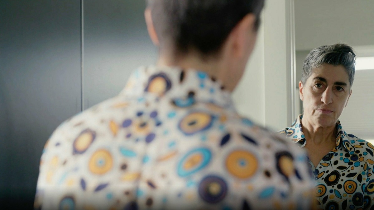 Sarah looks at herself in the mirror. She’s wearing a colorful shirt with designs in blue, orange, and purple.