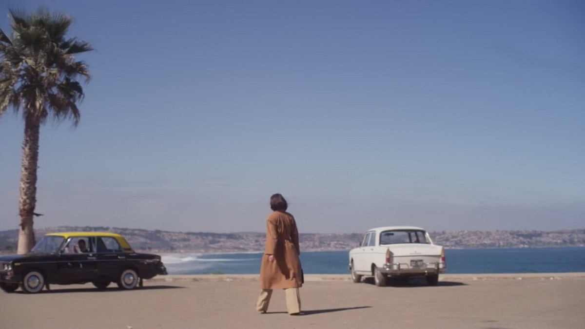 A  woman wearing beige trousers and a trench coat is walking towards one of the two cars parked on the beach.
