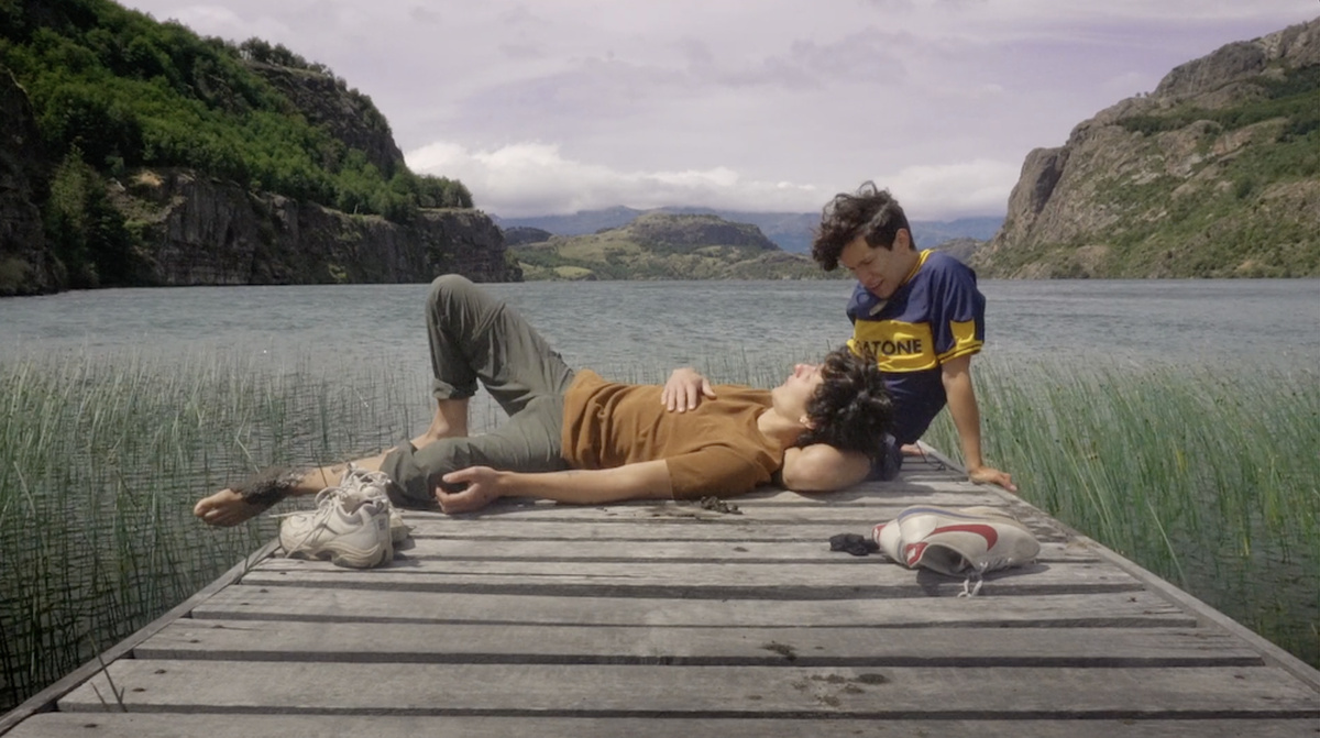 Two young men wearing colourful Tshirts on a wooden deck overlooking a lake; one is resting his head in the other's lap.