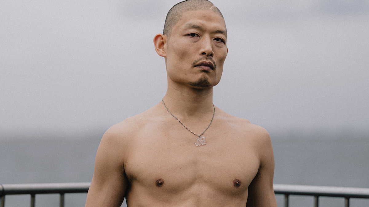 A shirtless Asian man stands in focus against the background of the sea, wearing a pendant necklace that says 'No War'.