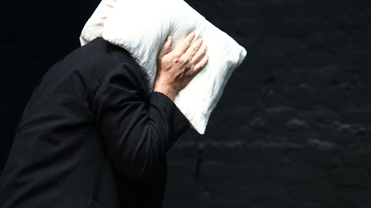 A person in a black jacket wraps a white pillow around their head and ears in front of a black background.
