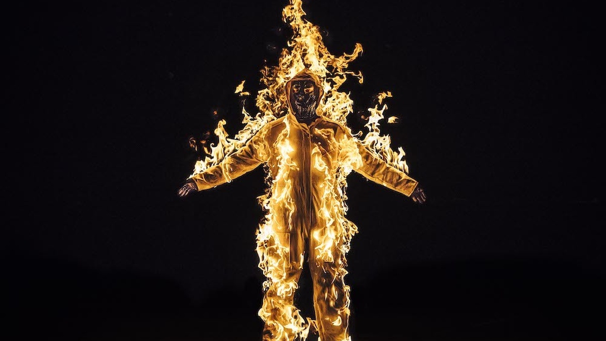 A masked and full body-covered figure, burning all over in a lit fire on their body, stands against a black background.