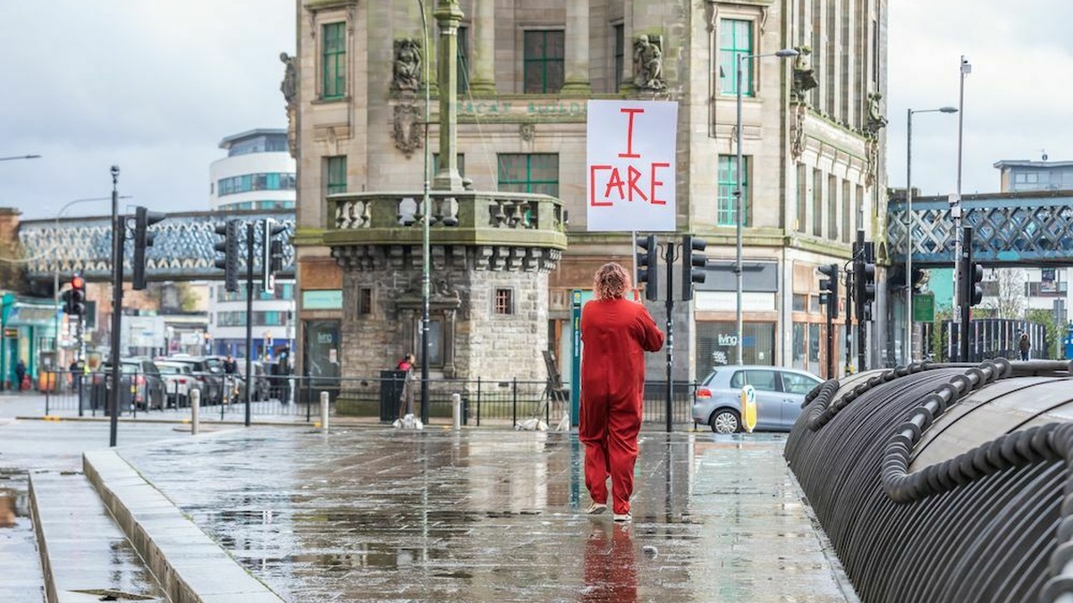 A person dressed in red, holding a placard with the words 'I care', in a Glasgow street, daytime.