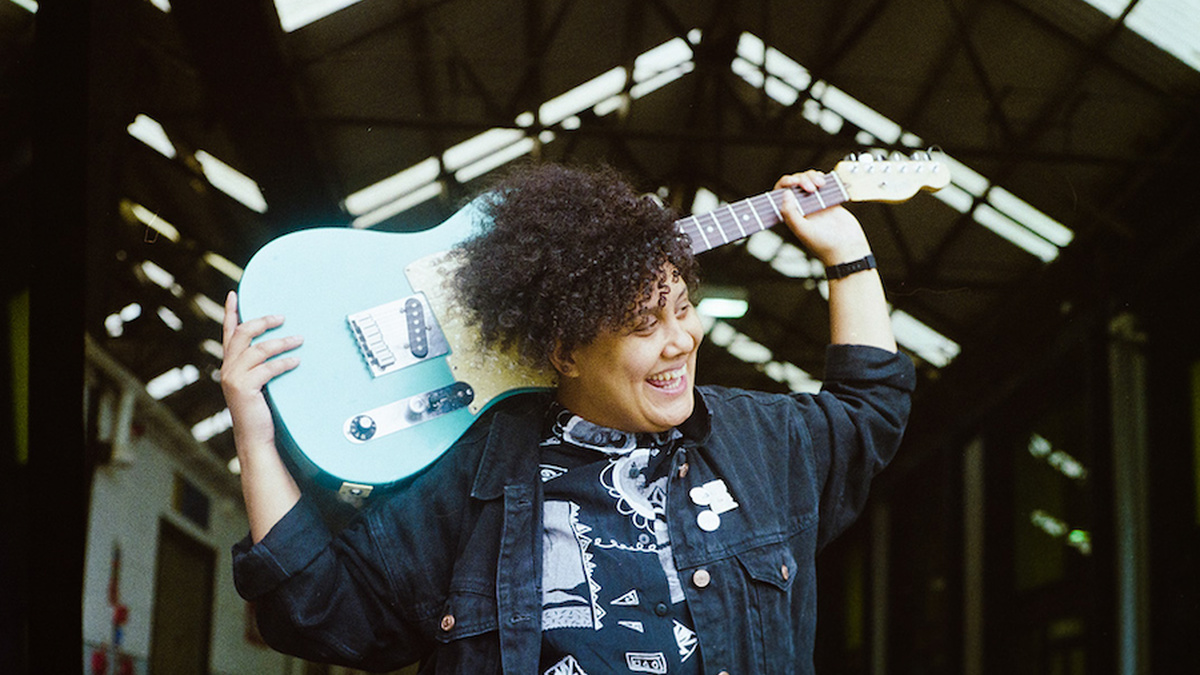Ray Aggs is pictured smiling, holding an electric guitar on their shoulders.