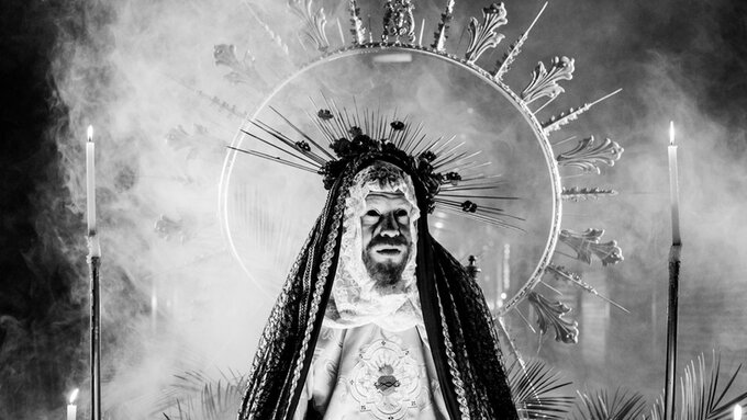 Black and white photograph of a man dressed as the Virgin Mary. There are candles, smoke and plants.
