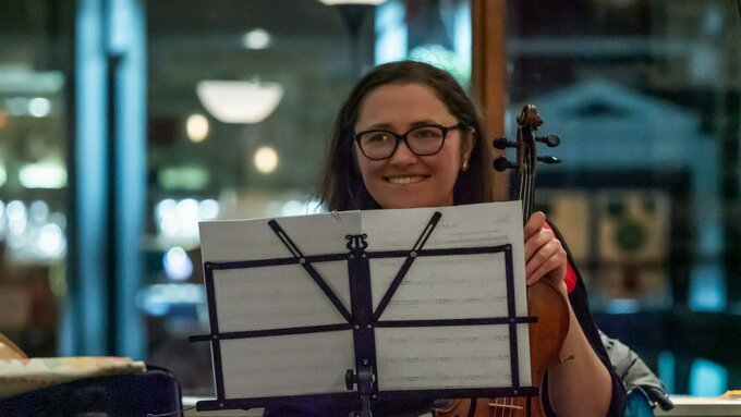 A white person with long dark hair wearing glasses, at a CoMA rehearsal holding a violin