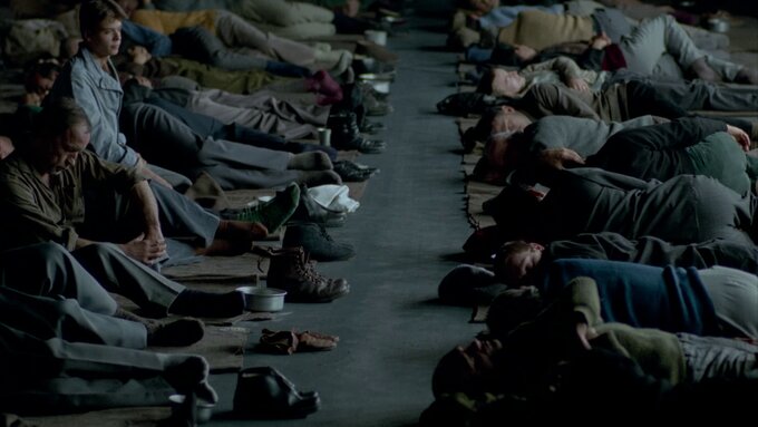 Two rows of people sleeping on the floor. They are dimly lit and they have no pillows or covers. Two men are sitting.