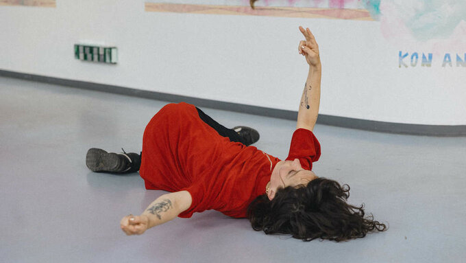 A white person with long dark hair in a red dress, they on the floor with their arm in the air making a peace sign.