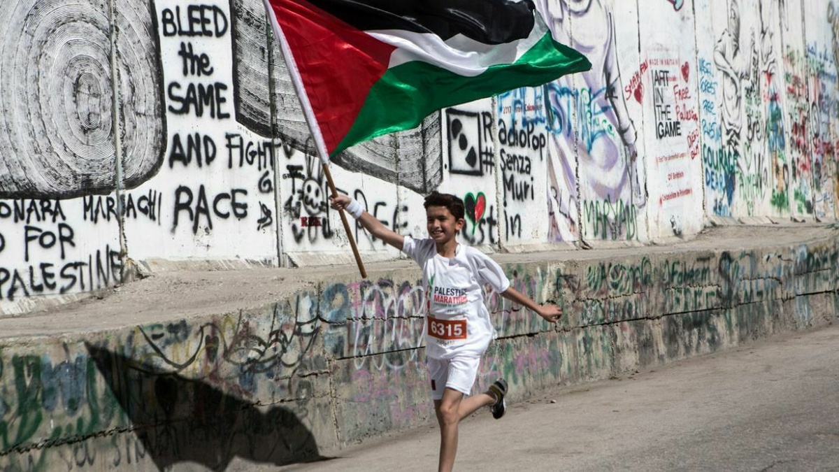 A young boy wearing running shorts and t-shirt, runs beside a high wall carring a Palestinian flag.