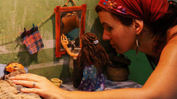 A person with a headscarf on the right leans into a clay animation scene of a little girl looking in the mirror