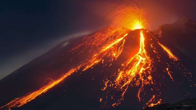 An erupting volcano at night, bright lava spouting from the top and flowing down the sides, against a dark blue sky.
