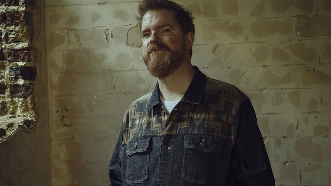 A photo of John Smith, a bearded white man wearing a denim button down stood in a disused industrial space