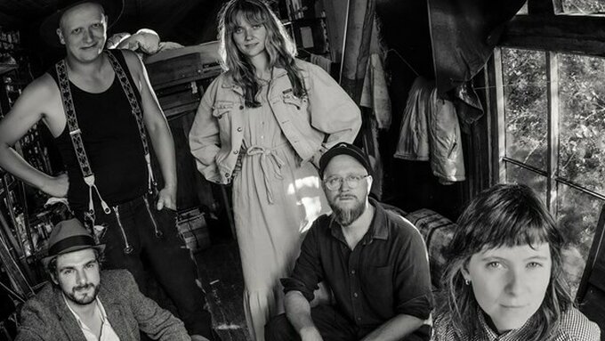 A black and white photo of 3 men and 2 women stood in an enclosed space. They are dressed in an indie-folk style.
