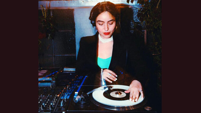 A femme person in a suit jacket, indigo top, wearing makeup. They are spinning a record on a DJ set.