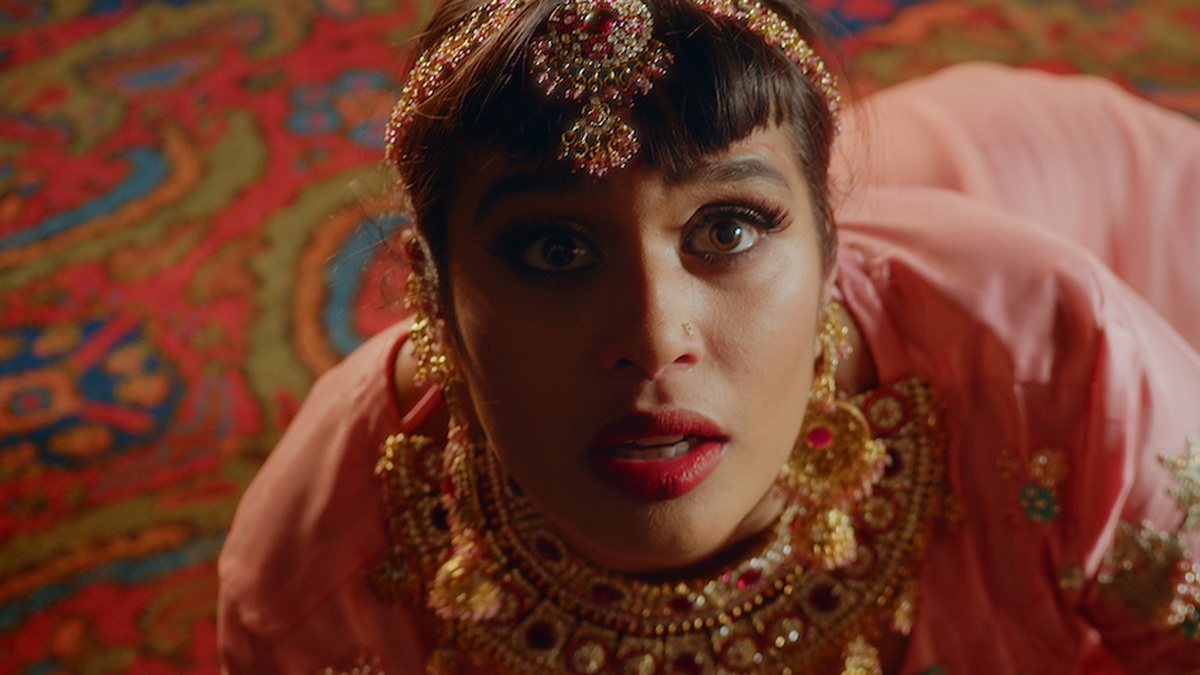 A brown skinned woman crouched facing the camera with a startled expression, she wears bright red lipstick and jewellery