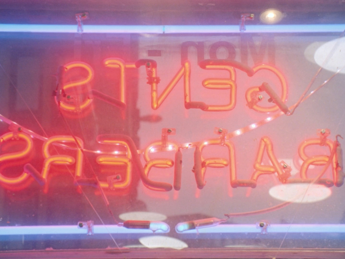 An orange neon sign in a shop window seen from behind, mirrored it reads "gents barbers".