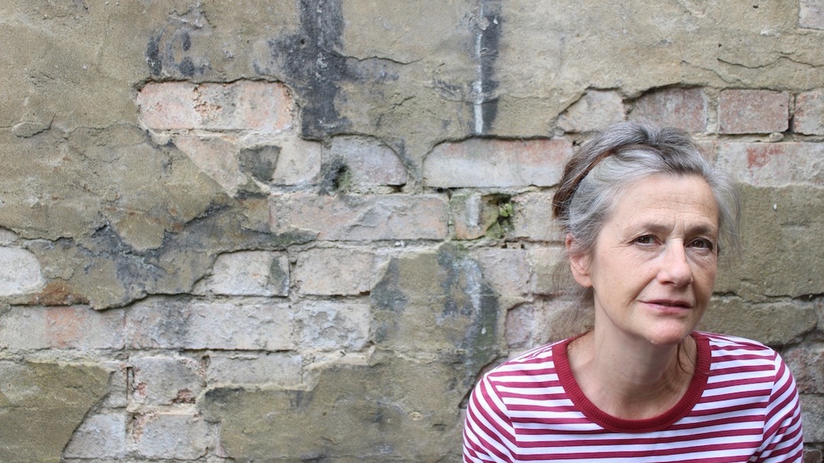 An older white woman with graying hair and dark brown eyes and is smiling slightly. She is stood in front of an old wall
