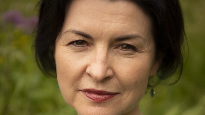A middle aged woman with dark brown centre-parted hair and narrow features against a green grassy background
