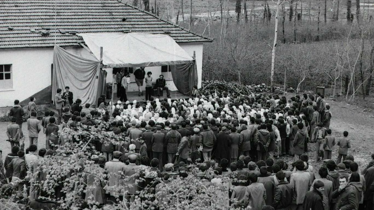 B&W photo of a mass of people attending a political meeting - in the background we see speakers on an makeshift stage