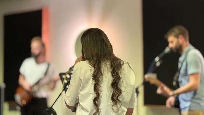 A photo centred on a person with long hair facing away from the camera, with 2 out of focus guitarists in the background