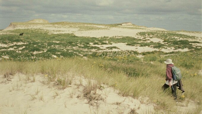 A still from 'Geographies of Solitude'. A vast island landscape. A person in a white sunhat walks through a dune.