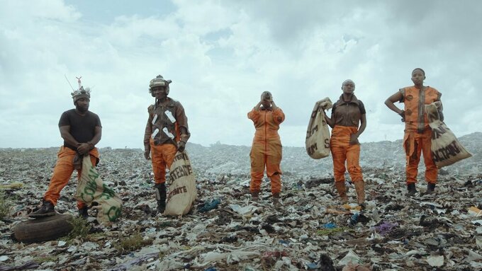 Five people stand on a landscape covered in rubbish. They are each wearing orange workwear, and carry hessian sacks.