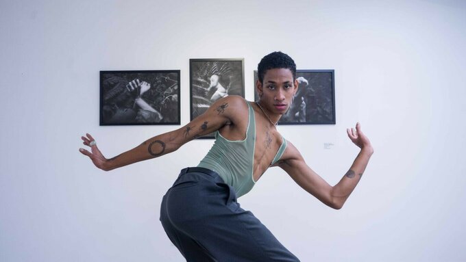 A dancer with tattoos and tight clothing poses in front of the camera. Behind them are three black and white photos.